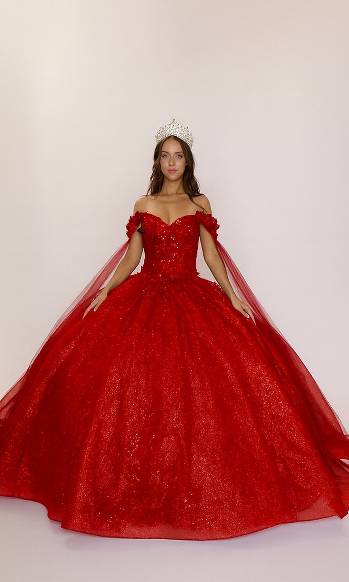 A woman stands against a plain, light background in the Quinceanera Dress Q127 by Nina Canacci, featuring an ornate red design with an off-the-shoulder neckline and glitter tulle skirt, accessorized with a jeweled crown.